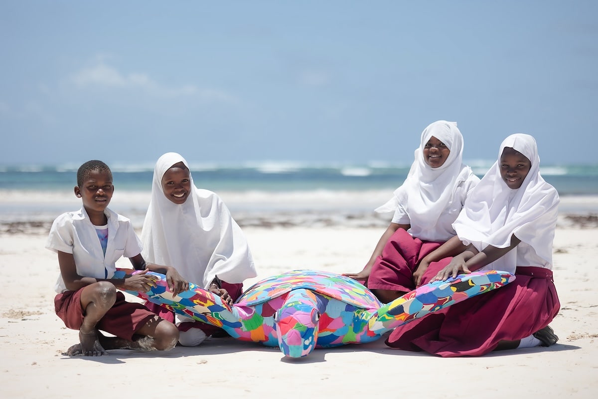 Four women sitting on a beach with a large turtle sculpture made of discarded and upcycled flip-flips